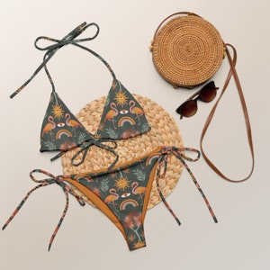 May include: A green and orange bikini with a flamingo and rainbow print. The bikini top is a triangle style and the bottom is a tie-side style. The bikini is on a woven straw mat with a brown wicker bag and sunglasses.