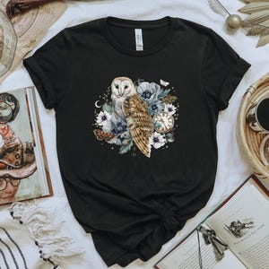 May include: Black t-shirt with a white barn owl illustration surrounded by flowers and a pocket watch. The owl has brown and white feathers and is perched on a branch with flowers.