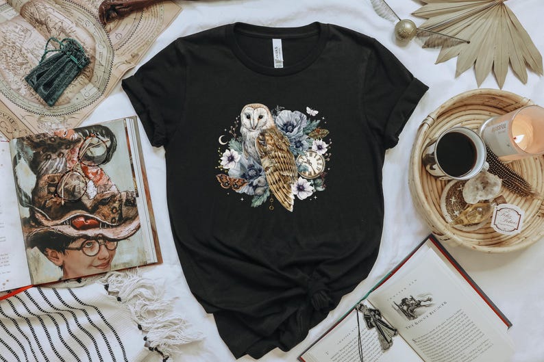 May include: Black t-shirt with a white barn owl illustration surrounded by flowers and a pocket watch. The owl has brown and white feathers and is perched on a branch with flowers.