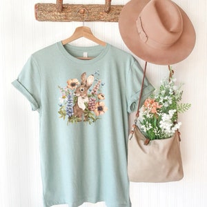 Op de afbeelding: Een lichtblauw t-shirt met een afbeelding van een bruin konijn omringd door kleurrijke bloemen. Het shirt hangt aan een houten rek met een bruine hoed en een bruine tas met bloemen.