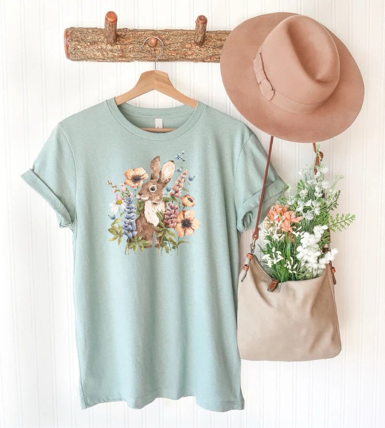 Op de afbeelding: Een lichtblauw t-shirt met een afbeelding van een bruin konijn omringd door kleurrijke bloemen. Het shirt hangt aan een houten rek met een bruine hoed en een bruine tas met bloemen.