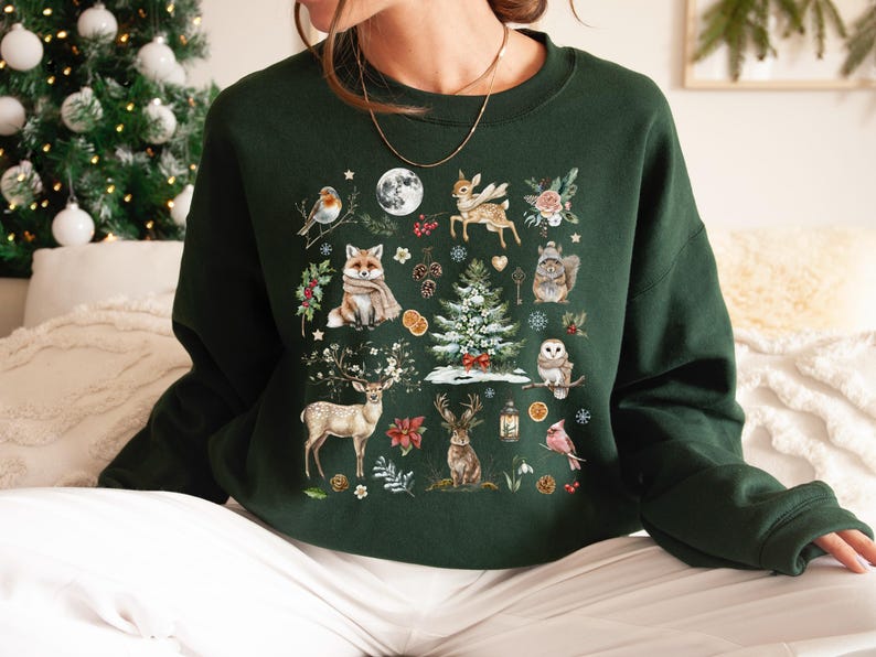 May include: A dark green sweatshirt with a festive winter scene printed on the front. The design features a variety of animals, including a fox, deer, and a bird, along with a Christmas tree and other wintery elements.