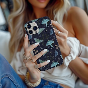 Puede incluir: Una funda de teléfono negra con un patrón de polilla azul oscuro y verde. La funda tiene un recorte para la lente de la cámara y un diseño de luna creciente blanca.