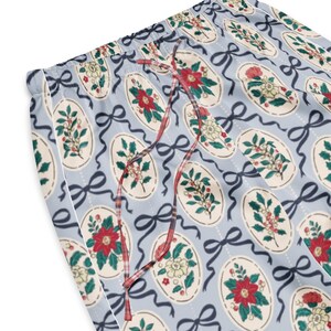 May include: Light blue pajama pants with a repeating pattern of floral ovals, dark blue bows, and a red and white plaid drawstring. The floral designs include red poinsettias, white flowers, and green leaves. White piping trims the edge of the pants.