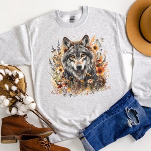 May include: A light grey crewneck sweatshirt featuring a central graphic of a wolf surrounded by orange and yellow wildflowers. The design includes detailed fur textures and botanical elements in warm autumn tones printed on the front of the garment.