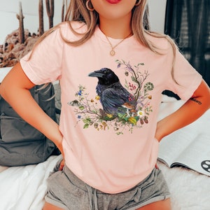 May include: A pink t-shirt with a black raven perched on a branch with colorful flowers and leaves. The raven is surrounded by a variety of natural elements, including mushrooms, acorns, and berries.