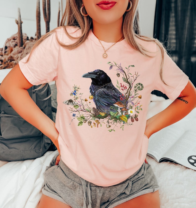 May include: A pink t-shirt with a black raven perched on a branch with colorful flowers and leaves. The raven is surrounded by a variety of natural elements, including mushrooms, acorns, and berries.