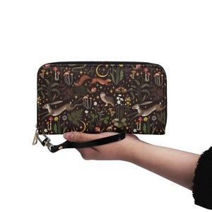 May include: A dark brown zippered wallet with a whimsical forest pattern featuring hares, foxes, mushrooms, and crescent moons. The wallet has a black wrist strap and a gold-coloured zipper pull.