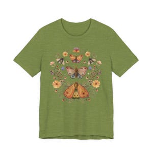 May include: An olive green short-sleeve t-shirt featuring a central graphic print of three butterflies and moths surrounded by various orange and yellow flowers, small green leaves, and tiny sparkling star accents arranged in a circular pattern.
