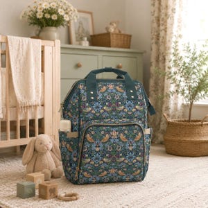 Op de afbeelding: Een luiertas met bloemenpatroon in blauw, groen en beige tinten. De rugzak heeft meerdere vakken, een handvat aan de bovenkant en een flessenhouder. De tas staat op een lichtgekleurd tapijt.