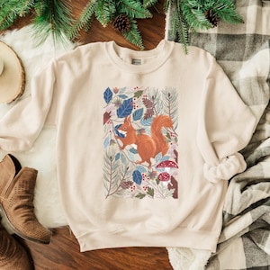 May include: A beige sweatshirt featuring a graphic print of a squirrel surrounded by leaves and flowers. The squirrel is orange and brown, and the leaves are blue, green, and red.