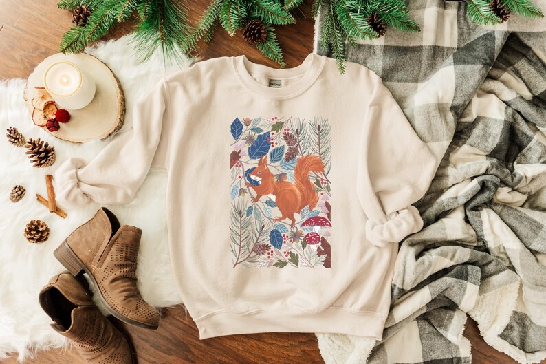 May include: A beige sweatshirt featuring a graphic print of a squirrel surrounded by leaves and flowers. The squirrel is orange and brown, and the leaves are blue, green, and red.