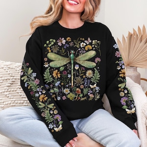 Può includere: Felpa nera con un design di libellula circondata da fiori colorati, farfalle e funghi. Le maniche sono decorate con motivi floreali. Il design ha un effetto acquerello.