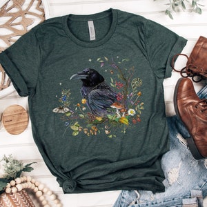 May include: A dark green t-shirt with a black raven surrounded by flowers, mushrooms, and leaves. The raven is perched on a branch and has a detailed design.