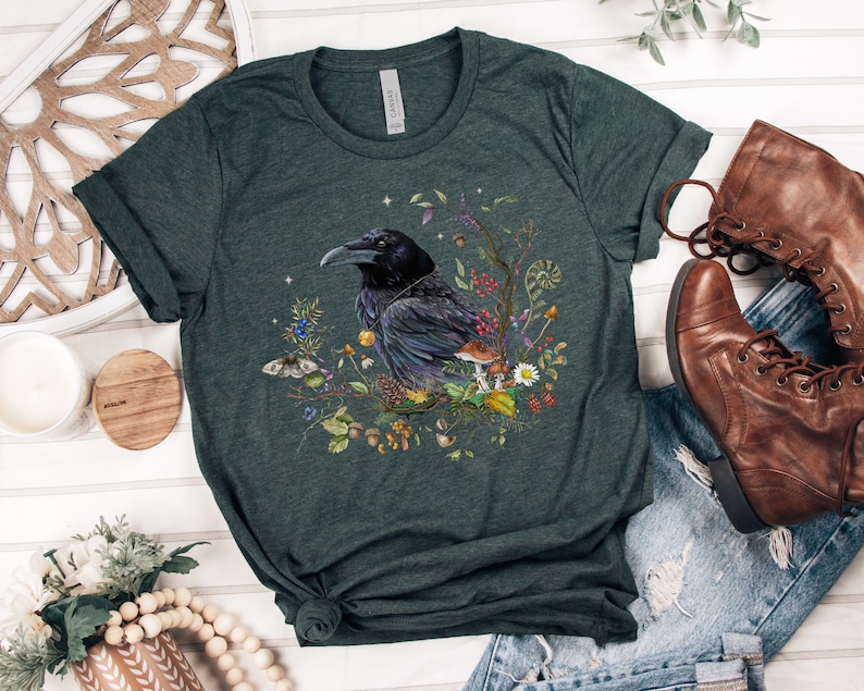 May include: A dark green t-shirt with a black raven surrounded by flowers, mushrooms, and leaves. The raven is perched on a branch and has a detailed design.