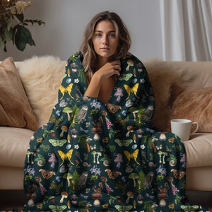 May include: A dark green throw blanket featuring a repeating pattern of mushrooms, butterflies, owls, and other woodland elements. The blanket is draped over a sofa and a person is sitting on the sofa.