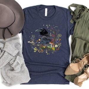 May include: A navy blue t-shirt with a graphic of a black raven surrounded by flowers and mushrooms. The raven is perched on a branch with its wings spread. The flowers are in shades of pink, purple, yellow, and white. The mushrooms are brown and white.