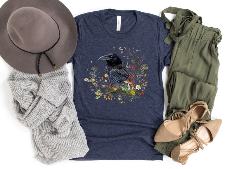 May include: A navy blue t-shirt with a graphic of a black raven surrounded by flowers and mushrooms. The raven is perched on a branch with its wings spread. The flowers are in shades of pink, purple, yellow, and white. The mushrooms are brown and white.