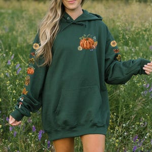 Puede incluir: Una sudadera con capucha de gran tama&ntilde;o de color verde oscuro con un dise&ntilde;o bordado de calabaza y follaje de oto&ntilde;o en la parte delantera. La sudadera tiene una capucha con cord&oacute;n y un bolsillo canguro.