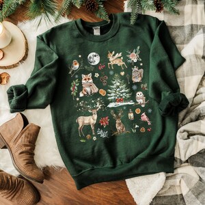 May include: A green sweatshirt with a festive winter scene featuring a fox, deer, owl, and other woodland creatures. The design is printed on a dark green background.