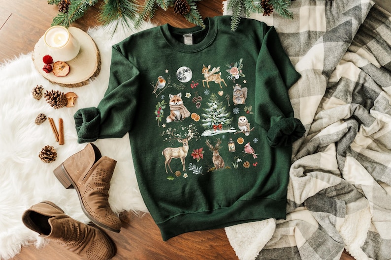 May include: A green sweatshirt with a festive winter scene featuring a fox, deer, owl, and other woodland creatures. The design is printed on a dark green background.