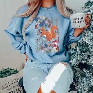 May include: A light blue sweatshirt featuring a graphic of a squirrel in a woodland scene. The text "Sweater Weather" is on a mug in the image.