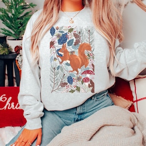 May include: A grey sweatshirt featuring a colourful illustration of a squirrel in a woodland setting. The squirrel is brown with a bushy tail. The woodland scene includes leaves, pine cones and mushrooms.