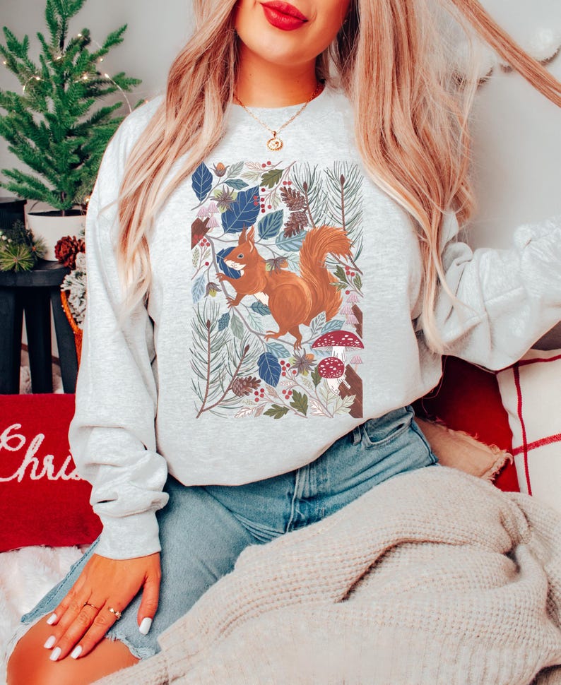 May include: A grey sweatshirt featuring a colourful illustration of a squirrel in a woodland setting. The squirrel is brown with a bushy tail. The woodland scene includes leaves, pine cones and mushrooms.
