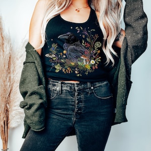K&ouml;nnte beinhalten: Ein schwarzes Tanktop mit einem grafischen Aufdruck eines Rabens, umgeben von Blumen und Bl&auml;ttern. Der Rabe sitzt auf einem Ast und hat ein dunkles, fast schwarzes Gefieder. Die Blumen sind in Schattierungen von Rosa, Lila und Wei&szlig;. Das Laub ist gr&uuml;n und braun.