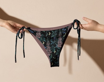 Op de afbeelding: Een zwarte en paarse bikini slip met een bloemenprint en verstelbare bandjes. De bikini slip heeft een lage taille en een cheeky cut.