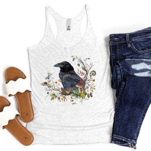 K&ouml;nnte beinhalten: Ein wei&szlig;es Tanktop mit einer schwarzen Rabenillustration, umgeben von Blumen, Bl&auml;ttern und Beeren. Der Rabe sitzt auf einem Ast und hat ein detailliertes Design. Das Tanktop ist in der Farbe Grau meliert und hat einen Racerback-Stil.