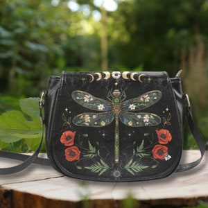 May include: Black leather crossbody bag with a dragonfly design. The dragonfly has floral details and is surrounded by poppies and ferns. The bag has a gold moon and eye design at the top.