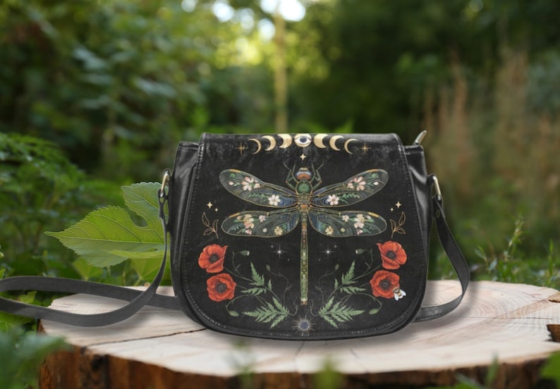 May include: Black leather crossbody bag with a dragonfly design. The dragonfly has floral details and is surrounded by poppies and ferns. The bag has a gold moon and eye design at the top.