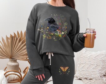 May include: Dark gray sweatshirt and sweatpants set. The sweatshirt features a detailed illustration of a raven surrounded by flowers. The sweatpants have a butterfly design. The set is made of a soft material.