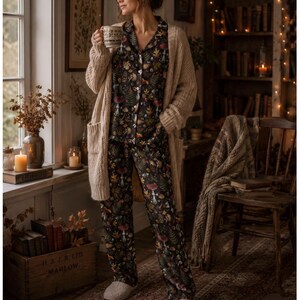 Puede incluir: Una mujer con un cárdigan beige sobre un pijama floral oscuro, sosteniendo una taza. El pijama presenta un estampado botánico con setas y follaje. Lleva zapatillas beige. La escena se desarrolla en una habitación acogedora con una estantería de madera.