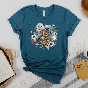 May include: Teal blue t-shirt with a white barn owl illustration surrounded by flowers and a pocket watch. The owl is perched on a branch with its wings spread. The flowers are white and pink with green leaves. The pocket watch is gold with Roman numerals.