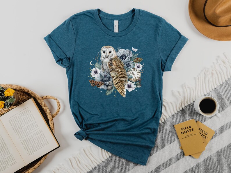 May include: Teal blue t-shirt with a white barn owl illustration surrounded by flowers and a pocket watch. The owl is perched on a branch with its wings spread. The flowers are white and pink with green leaves. The pocket watch is gold with Roman numerals.