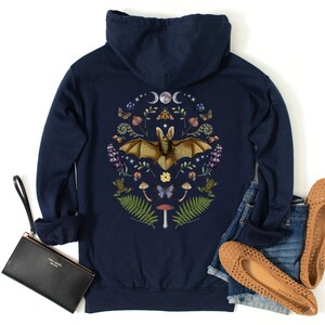 May include: Navy blue hooded sweatshirt featuring a graphic print of a bat surrounded by flowers, mushrooms, and other woodland elements. The graphic is in a circle with a crescent moon and stars above the bat.