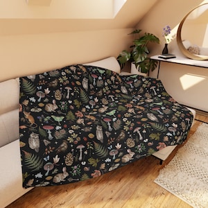 May include: A black throw blanket with a woodland pattern featuring mushrooms, owls, rabbits, and other forest creatures. The blanket is made of soft, comfortable fabric and is perfect for adding a touch of nature to your home.