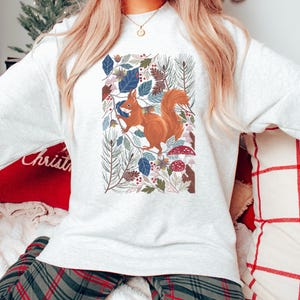 May include: A white sweatshirt featuring a printed design of a red squirrel surrounded by leaves, pine needles, and mushrooms.