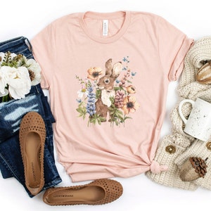 Op de afbeelding: Een roze T-shirt met een bruin konijn omringd door kleurrijke bloemen. Het shirt ligt op een witte achtergrond met een paar bruine sandalen, een blauwe jeans, een witte mok en een beige gebreide sjaal.