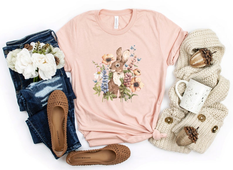 Op de afbeelding: Een roze T-shirt met een bruin konijn omringd door kleurrijke bloemen. Het shirt ligt op een witte achtergrond met een paar bruine sandalen, een blauwe jeans, een witte mok en een beige gebreide sjaal.