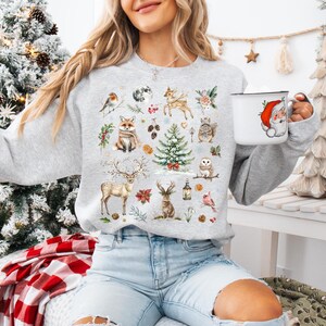 May include: A gray sweatshirt with a Christmas-themed design featuring a variety of woodland animals, including a fox, deer, owl, and birds. The design also includes a Christmas tree, pinecones, and other festive elements.