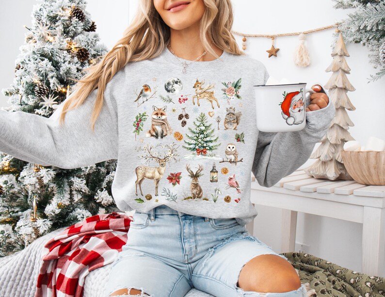 May include: A gray sweatshirt with a Christmas-themed design featuring a variety of woodland animals, including a fox, deer, owl, and birds. The design also includes a Christmas tree, pinecones, and other festive elements.