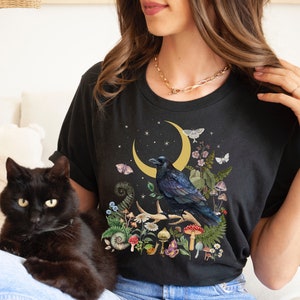 May include: A black t-shirt with a graphic of a raven perched on a branch with a crescent moon and stars in the background. The graphic also includes mushrooms, flowers, and other botanical elements. The t-shirt is being worn by a person with a black cat sitting on their lap.