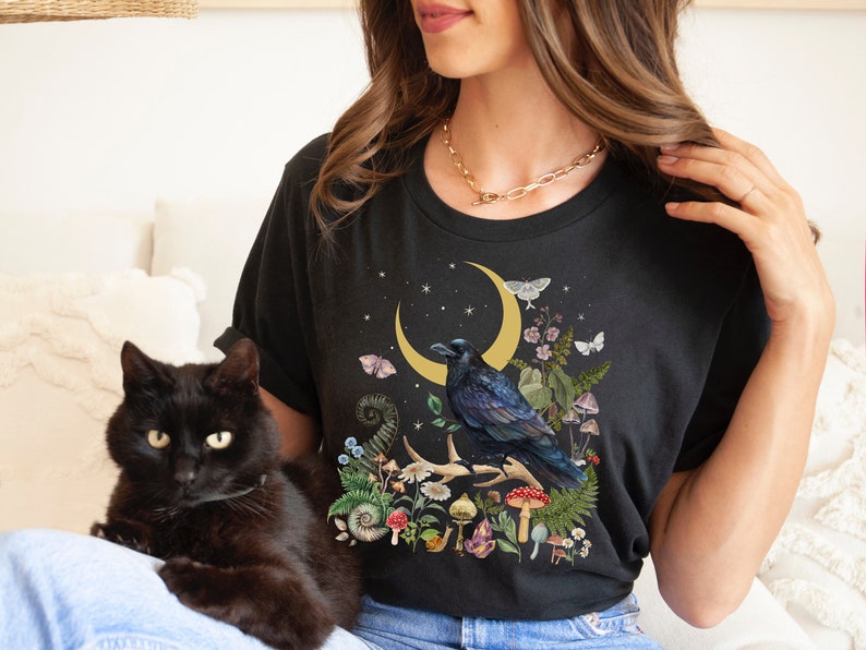 May include: A black t-shirt with a graphic of a raven perched on a branch with a crescent moon and stars in the background. The graphic also includes mushrooms, flowers, and other botanical elements. The t-shirt is being worn by a person with a black cat sitting on their lap.