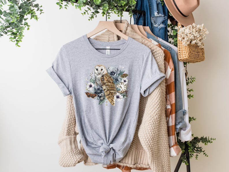 May include: A gray t-shirt with a white barn owl and floral design. The owl is brown and white with yellow eyes. The flowers are blue, white, and pink.