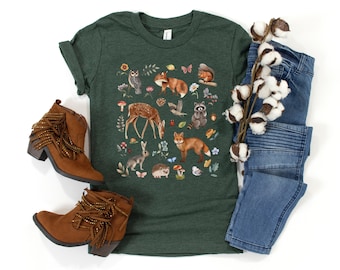 May include: A dark green short-sleeve t-shirt featuring a colorful graphic print of various woodland animals including a deer, foxes, a raccoon, a rabbit, a hedgehog, and an owl, surrounded by small flowers, mushrooms, and leaves.