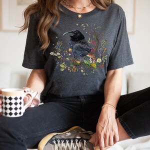 May include: A dark gray t-shirt with a graphic print of a black raven sitting in a nest of leaves, berries, and mushrooms. The raven has a detailed design with a gold beak and eyes.