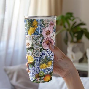 May include: A tall, insulated tumbler with a floral and fruit design. The design features yellow and pink flowers, oranges, kiwis, and blue and white tile patterns. The tumbler is held by a person.